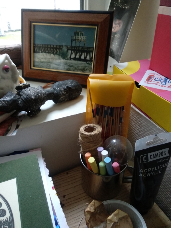 Une photo d'un coin de mon bureau en bazar. Dans un mug des mikados, de la ficelle, une ampoule et des craies. Un cadre avec une photo de la mer. Une statuette renversée et un chouette avec un googly-eye. Une pile de carnet, feuilles et un repose sachet de thé.