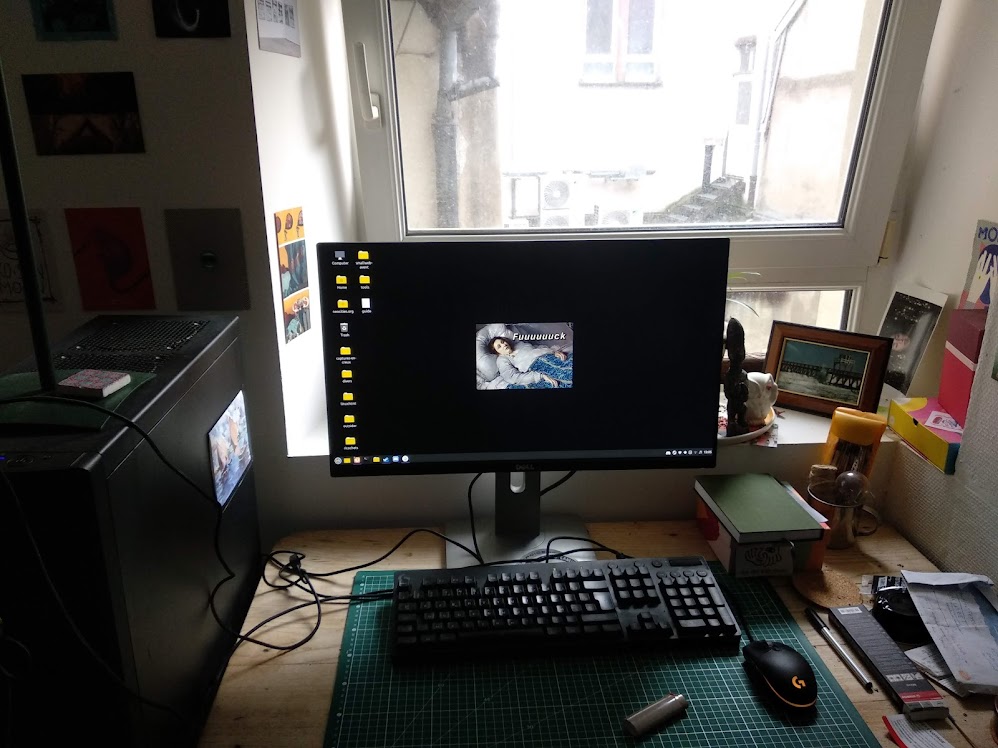 Mon atelier. Le clavier posé sur le plan à découpe. Des objets et des boîtes et des statuettes et des cartes postales au mur. Ce n'est pas très bien ordonné. Sur l'écran de l'ordinateur il y a en fond une peinture d'une femme dans son lit, difficile de lire l'expression de son visage mais a été ajouté un texte, Fuuuuuuuuck.